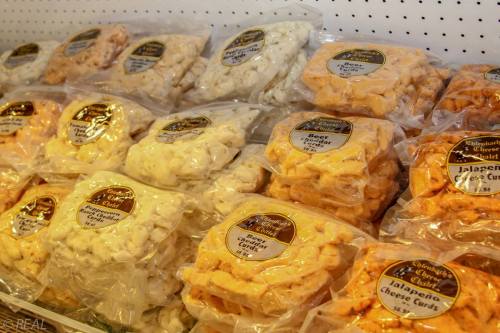 Bags of cheese curds in various flavors displayed on a shelf, labeled with product names and prices.