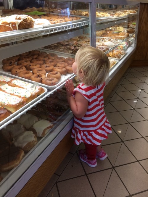 A small child in a striped dress gazes longingly at a display of pastries and donuts in a bakery.