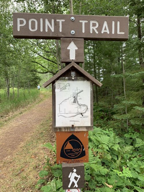 A wooden sign for the Point Trail, featuring a map and directional arrow, surrounded by greenery and a dirt path.