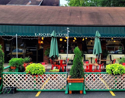 Outdoor dining area of a restaurant with green umbrellas, red chairs, and potted plants in front of a brick building.
