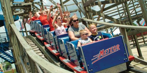 A group of excited people on a roller coaster, smiling and raising their hands, with a wooden structure in the background.