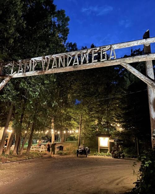 Wooden sign reading "Camp Wandawear" at night, surrounded by trees and string lights illuminating the path.