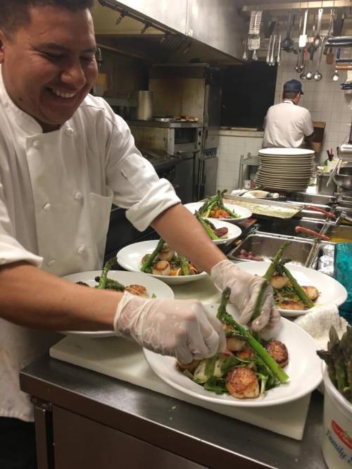 A chef in a kitchen plates scallops and asparagus, smiling as he prepares dishes for service.