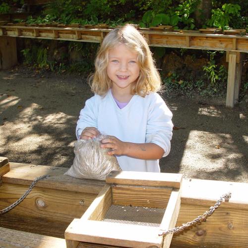 A smiling child with curly hair holds a small bag while standing near a wooden structure in a natural setting.
