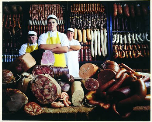 Three butchers stand proudly behind a display of various cured meats and sausages in a shop.