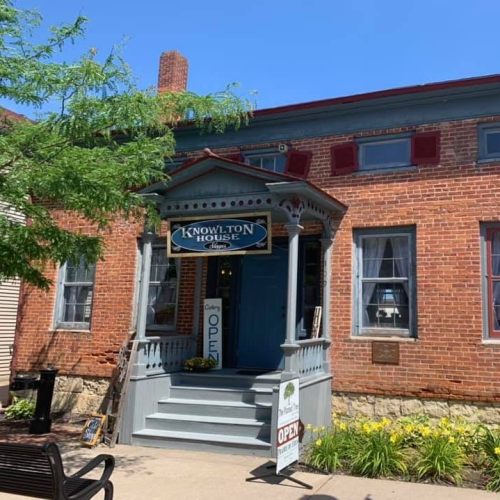 A brick building with a porch and sign reading "Knowlton House Gallery," surrounded by greenery on a sunny day.
