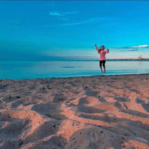 A person joyfully jumps on a sandy beach with calm water and a clear blue sky in the background.