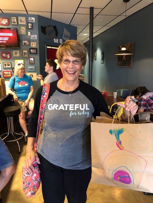 A smiling woman in a "Grateful for Today" shirt holds a colorful paper bag in a cafe setting.