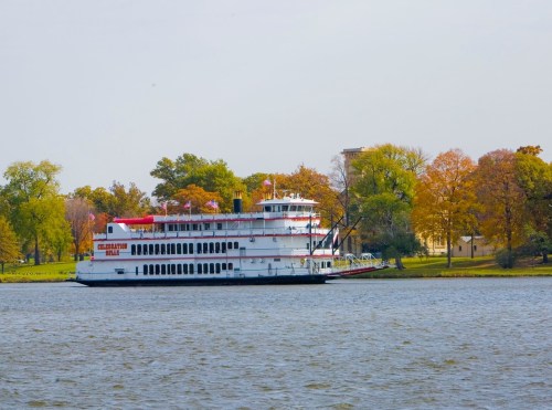 A large white riverboat with red accents sails on a calm lake, surrounded by trees with autumn foliage.