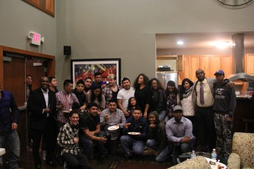 A diverse group of people gathered in a cozy indoor space, smiling and posing for a photo with food plates.