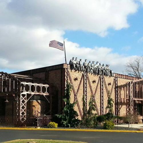 A brick building with a flagpole, featuring a group of statues on the roof and surrounded by trees and landscaping.