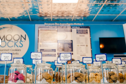 A display of gourmet cookies in jars, with labels for flavors like blueberry and maple chocolate chip, against a blue wall.