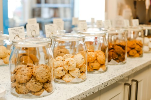 A display of various cookies in glass jars on a countertop, with labels in the background.