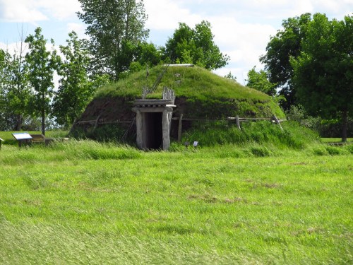 A grassy, dome-shaped structure with a wooden door, surrounded by trees and tall grass under a blue sky.