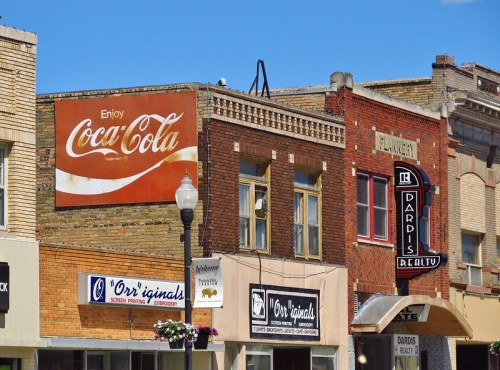 Vintage storefronts featuring a Coca-Cola sign and various local businesses under a clear blue sky.