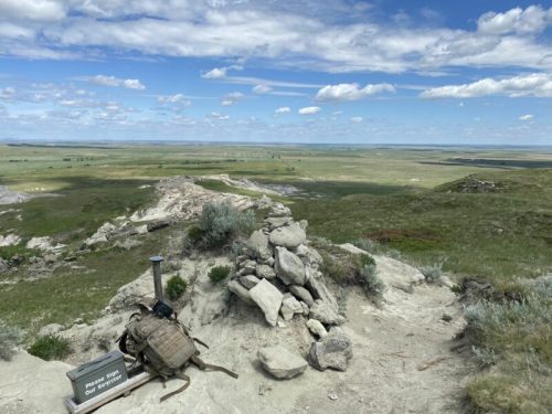 A rocky outcrop with a backpack and a sign, overlooking a vast green landscape under a blue sky with clouds.