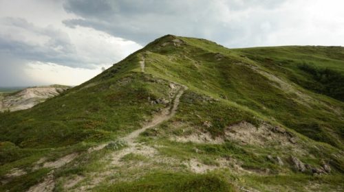 A winding path leads up a grassy hill under a cloudy sky, with rolling green landscapes in the background.