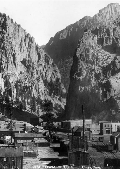 Black and white photo of Jim Town, Colorado, nestled between towering cliffs and mountains. Historic buildings line the valley.