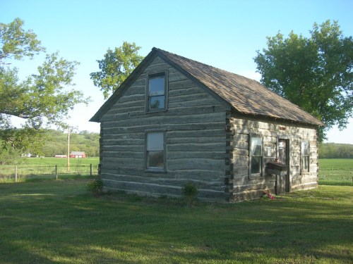 A rustic log cabin with a sloped roof, surrounded by green grass and trees, set against a rural landscape.