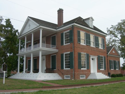 Historic brick house with white columns, green shutters, and a porch, set in a landscaped area.