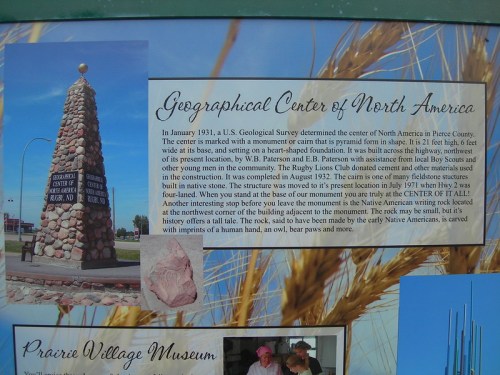 A stone monument marking the Geographical Center of North America, with informational text and a scenic background.