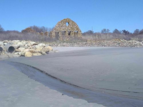 Ruins of a stone building on a sandy beach, with a clear blue sky and sparse vegetation in the background.