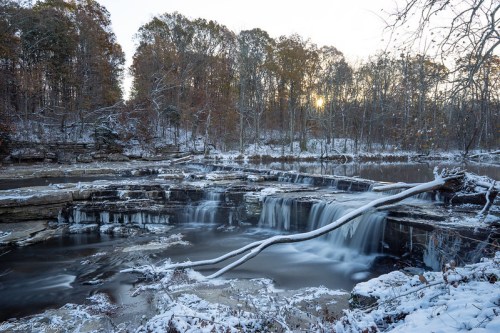 A serene winter scene featuring a waterfall surrounded by snow-covered rocks and trees, with a soft sunset glow.