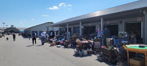 A bustling outdoor market with vendors selling various items, shoppers browsing, and clear blue skies above.
