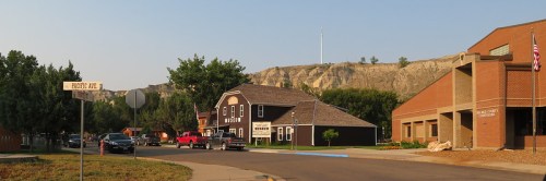 A small town street with a museum, public library, and rocky cliffs in the background under a clear sky.