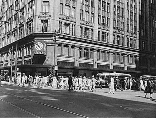 Black and white photo of a busy street scene with pedestrians and vintage buses in front of a large building.