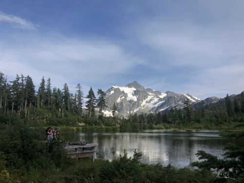 A serene mountain lake surrounded by trees, with snow-capped peaks in the background and people by the water's edge.