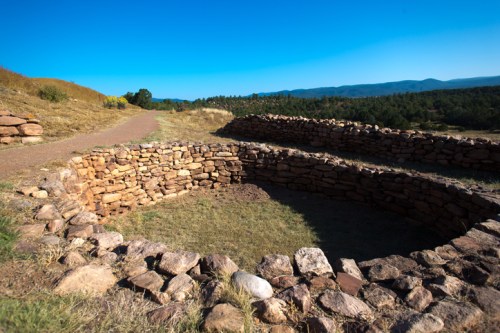 Stone ruins of circular structures on a grassy landscape, with rolling hills and a clear blue sky in the background.