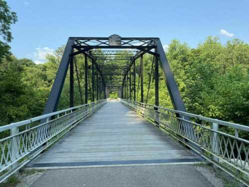 A metal truss bridge spans a path surrounded by lush green trees under a clear blue sky.