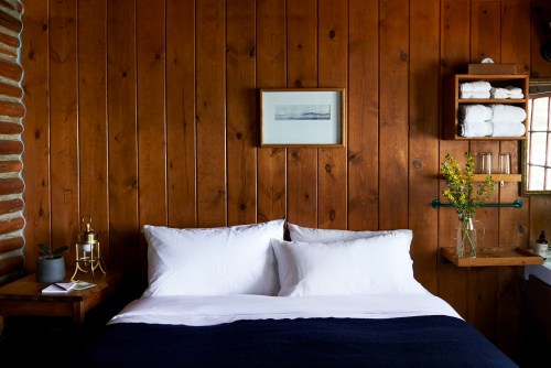 Cozy bedroom with wooden walls, a neatly made bed, and decorative shelves with towels and flowers.