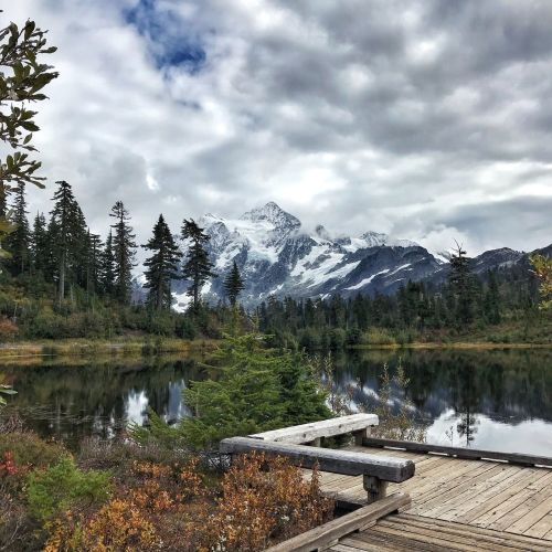Scenic view of a mountain landscape reflected in a calm lake, surrounded by lush trees and cloudy skies.