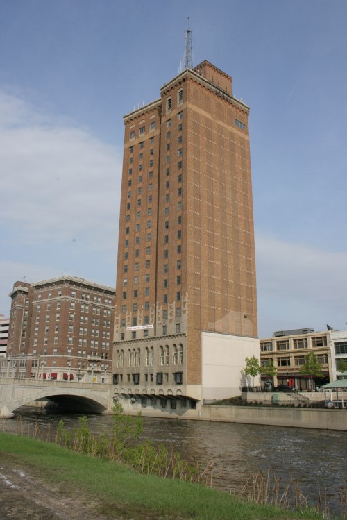 A tall brick building with a tower stands beside a river, featuring a bridge in the foreground and a clear sky above.