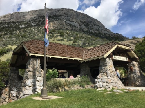 A stone visitor center with flags in front, set against a mountainous landscape under a blue sky with clouds.