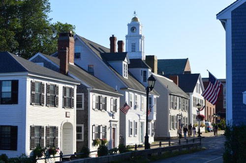 Historic buildings line a street, featuring American flags and a clock tower in the background under a clear blue sky.