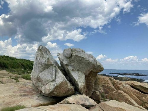 A large split rock on a coastal landscape under a cloudy sky, with the ocean visible in the background.