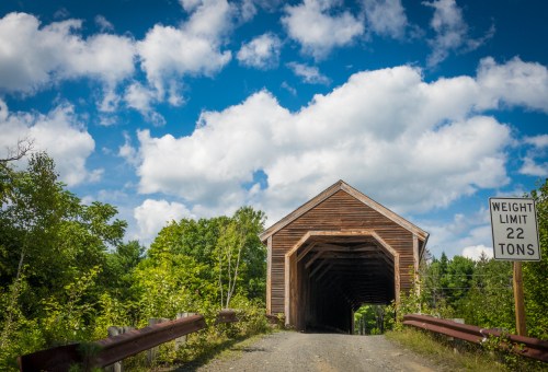 A wooden covered bridge leads into a lush green landscape under a bright blue sky with fluffy clouds.