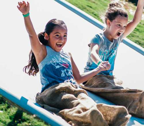 Two girls joyfully sliding down a hill on a sunny day, holding hands and laughing.