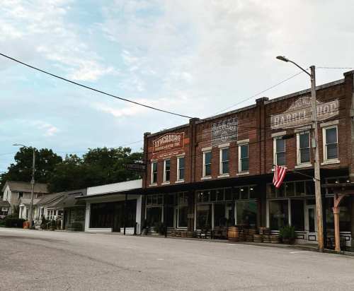 Historic storefronts line a quiet street, featuring vintage signage and an American flag, under a cloudy sky.