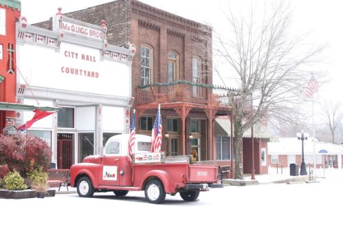 A vintage red truck parked in a snowy town square with historic buildings and flags in the background.