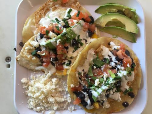 Two tacos topped with beans, tomatoes, avocado, sour cream, and cheese, served with a side of crumbled cheese.