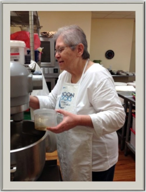 An older woman in an apron mixes ingredients in a kitchen, focused on her task with a bowl in hand.