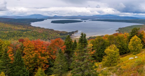 A scenic view of a lake surrounded by colorful autumn foliage and distant mountains under a cloudy sky.
