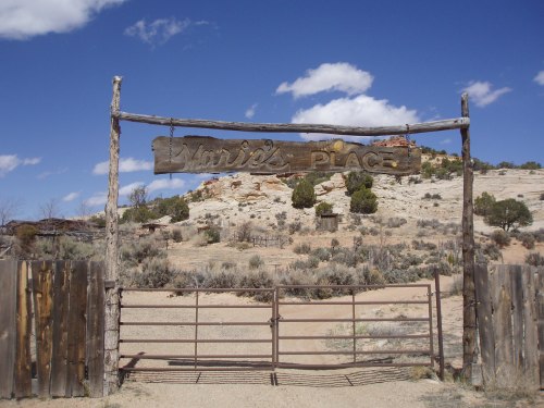 A rustic wooden sign reading "Marie's Place" hangs over a gate in a desert landscape with blue skies and sparse vegetation.