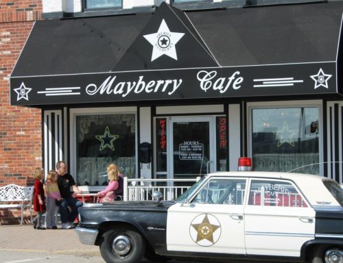 A vintage police car parked outside the Mayberry Cafe, with people sitting at a table nearby.