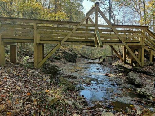 A wooden bridge spans a small stream in a forested area with autumn foliage and rocky terrain.