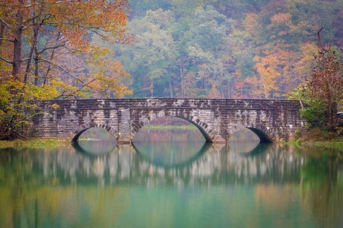 A stone bridge with arches spans a calm, reflective lake surrounded by colorful autumn trees.